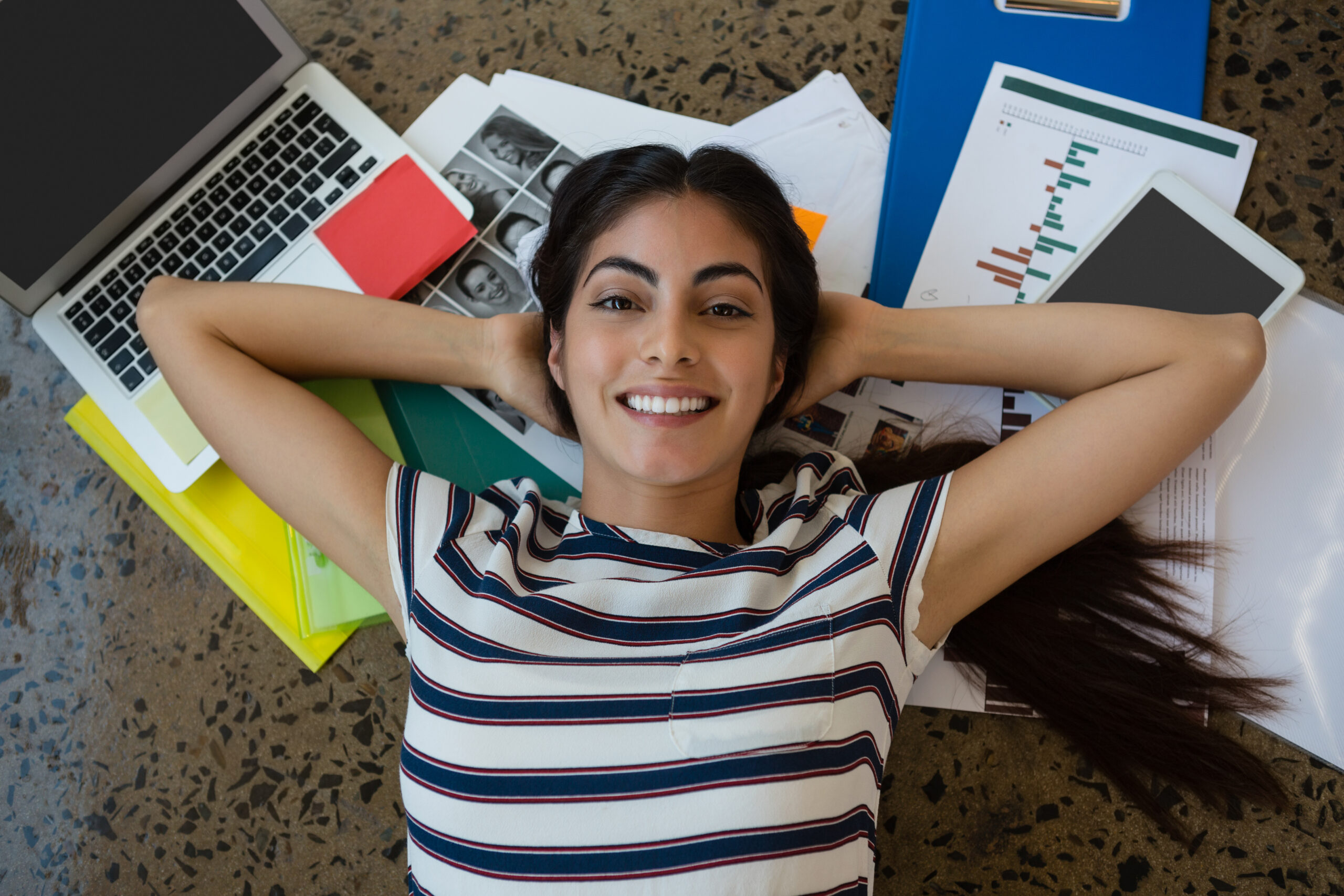 Portrait Of Smiling Beautiful Hispanic Woman Relaxing On Documents With Arms Behind Head At Creative Office